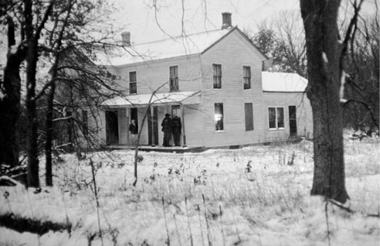 Imagen en blanco y negro de la granja de Ed Gein a los lejos, siendo registrada por la policía.